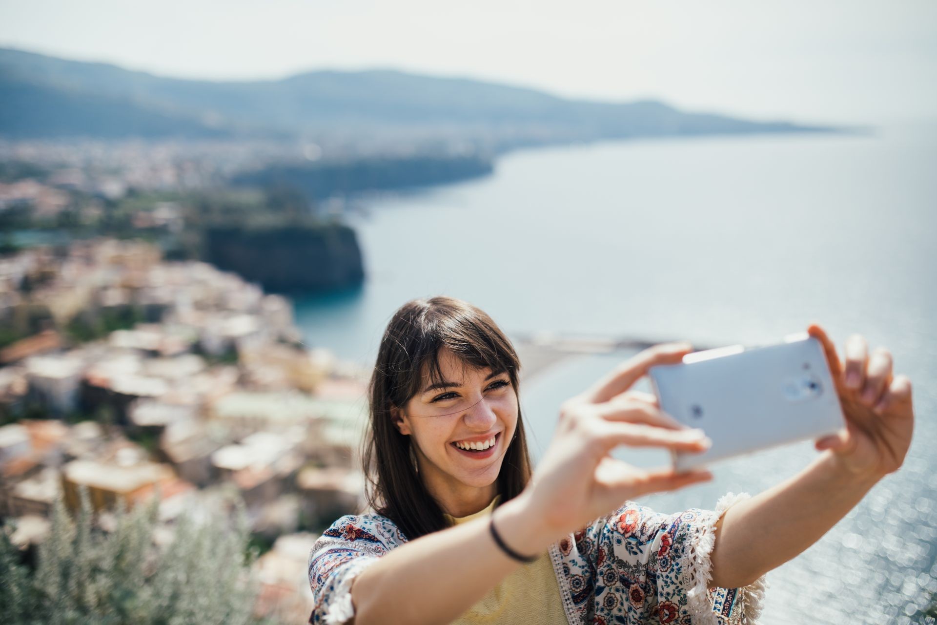 Happy woman traveler smiling at italian coast view.Woman traveling to European south coast.Enoying sunny weather mediterranean seaside.Taking selfie with smartphone front camera